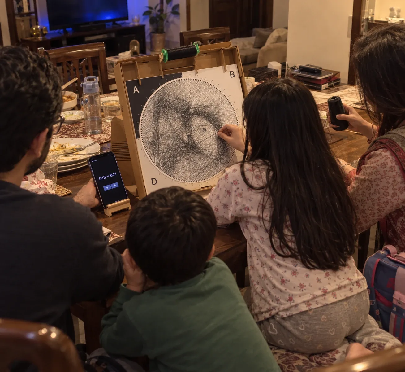 A family around the dining table after dinner, a girl weaving the portrait on the box stand while dad reads the step
